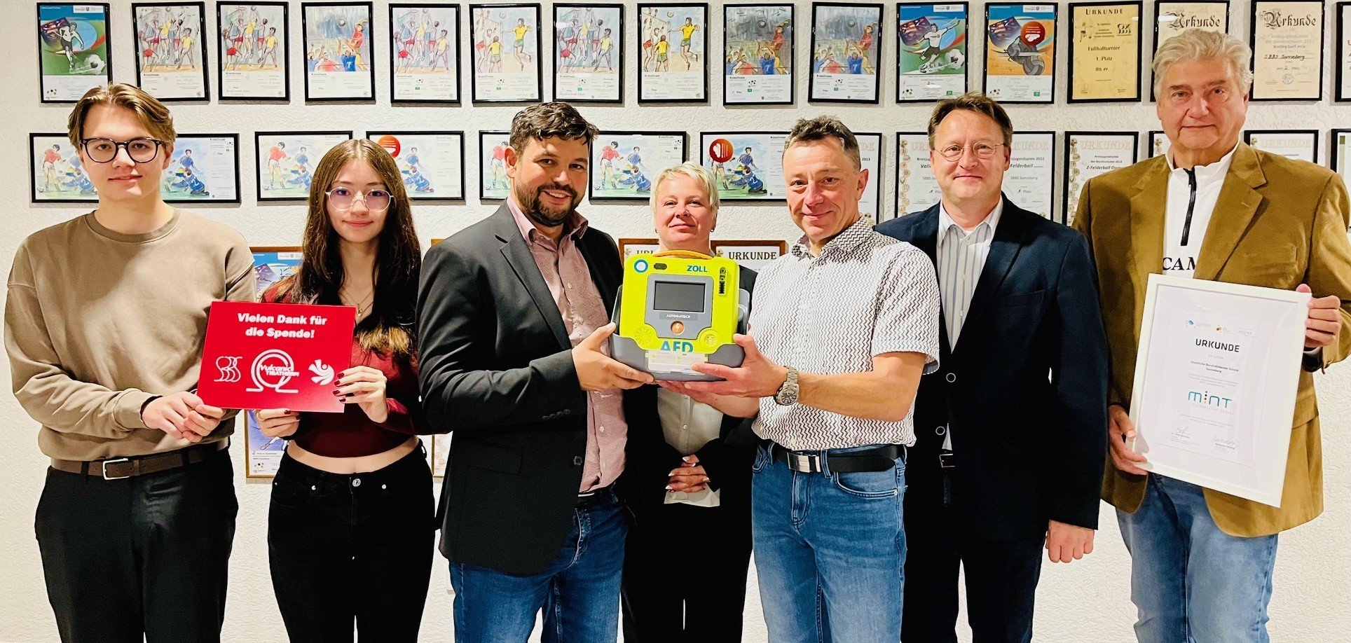 VULCANIC TRIATHERM-Geschäftsführer Andreas Fischer (3.v.l.) und Personalleiterin Anja Müller (M.) übergeben SBBS-Schulleiter Steffen Werner (3.v.r.) im Beisein von Landrat Robert Sesselmann (2.v.r.) den Defibrillator. Die Zwölftklässler Cedric Bauersachs (l.) und Angelina Bäz (2.v.l.) vom Beruflichen Gymnasium der SBBS zeigen die von ihnen gestaltete Hinweistafel zur Sachspende. Der stellvertretende SBBS-Schulleiter Uwe Blechschmidt (r.) hält die jüngst überreichte Urkunde „MINT-freundliche Schule“ in seinen Händen.